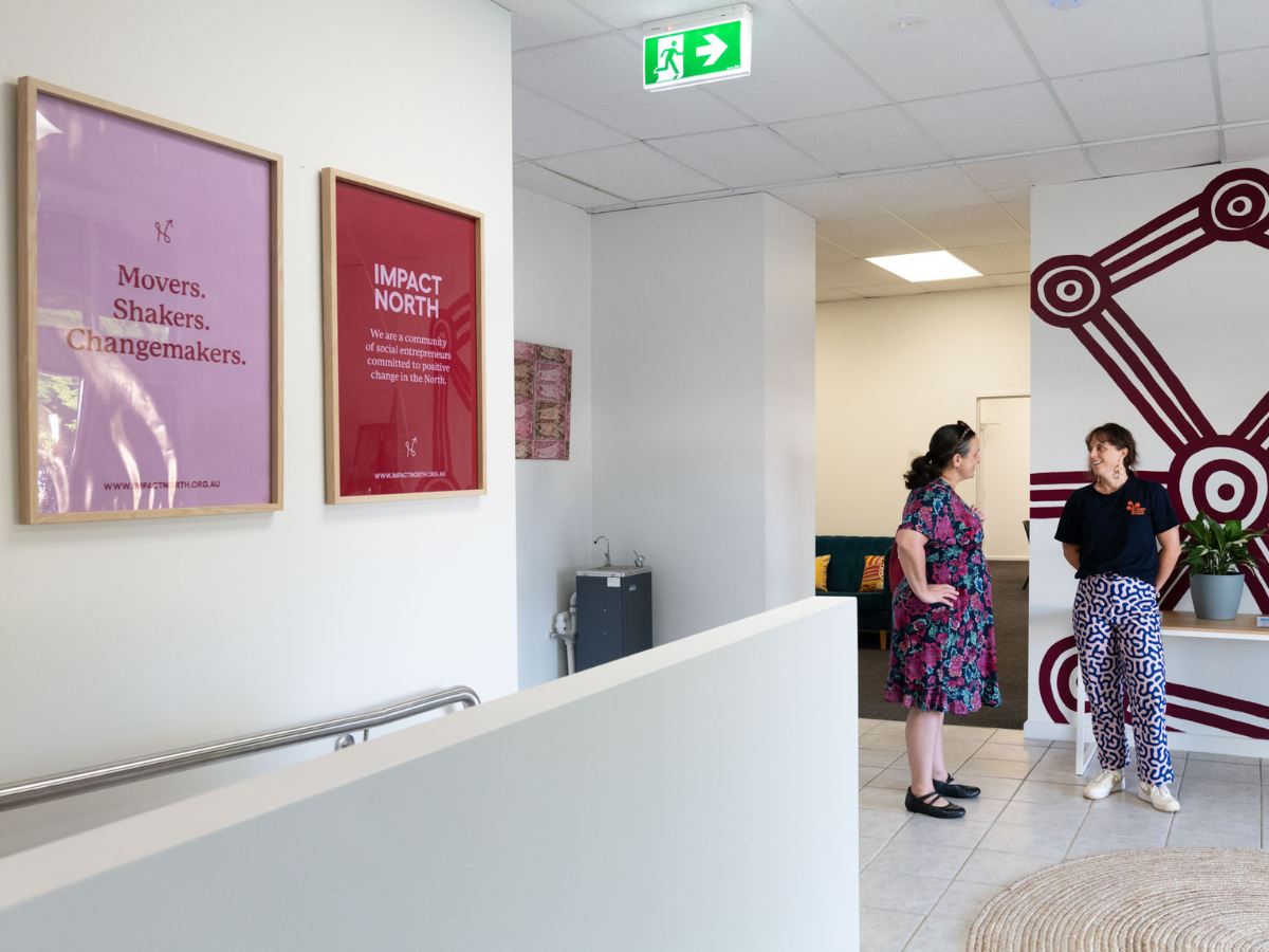 A room with white walls with artwork and picture frames containing inspiring quotes in pink and red in the foreground. Two women stand talking in the background.