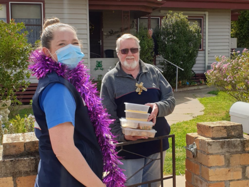 A woman delivers food to residents, handing it over a gate. Both are wearing Christmas tinsel.