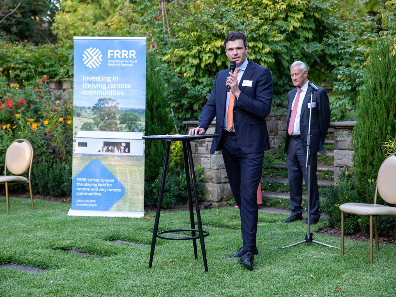 Man stands at a tall black table in a suit, talking into a microphone in a beautiful garden.