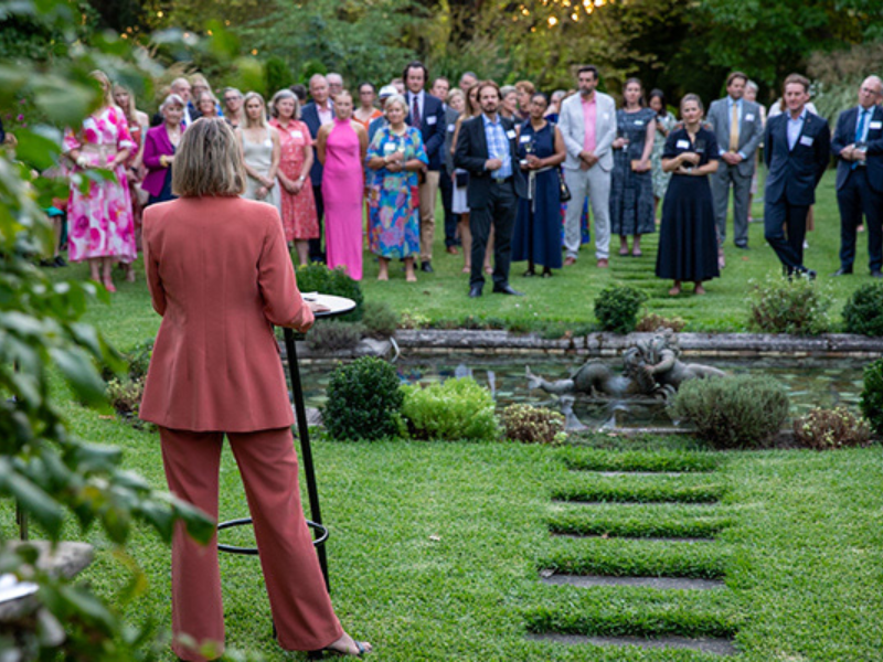 Image of woman wearing an orange suit talking to a group of guests, taken from behind her.