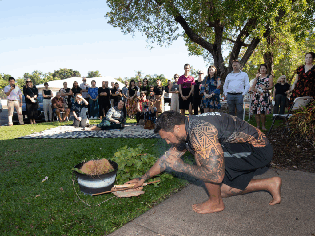 A man kneels on the floor in front of a small audience, performing a smoking ceremony.