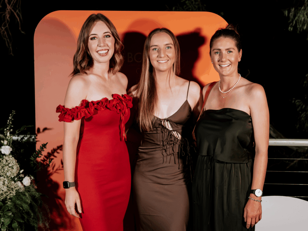 Three women are dressed formally and standing in front of a black and orange backdrop. 