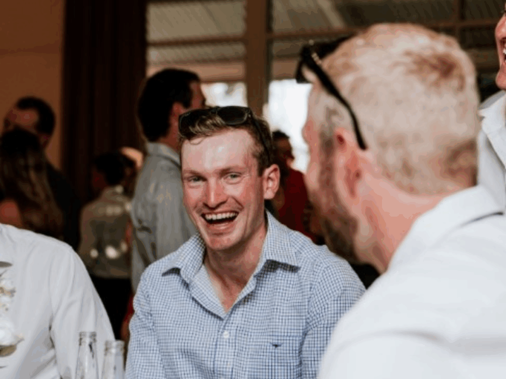 Image shows a close up of a man in a blue shirt widely smiling at the camera, with a blurred image of another man in the foreground. 
