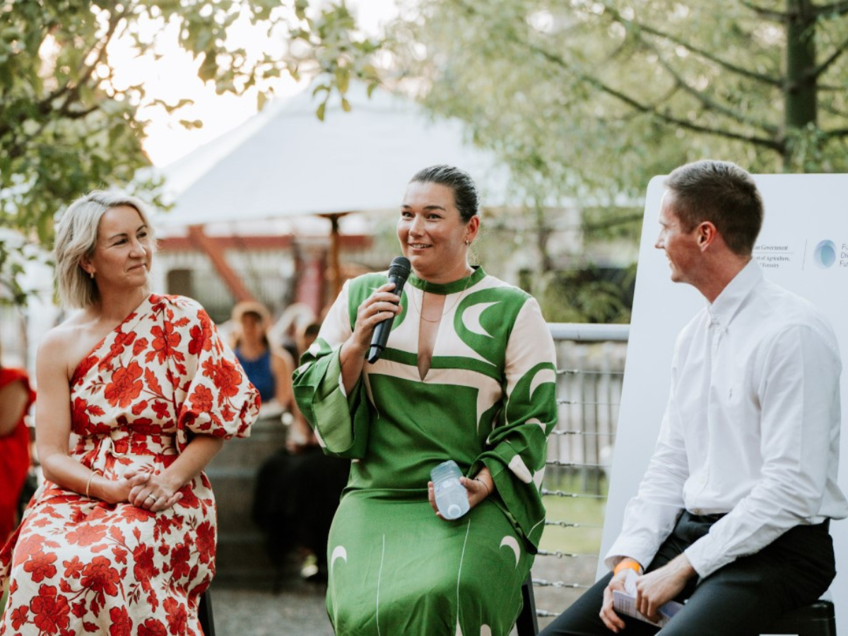 Two women and a man are dressed formally and speaking at an event. Middle woman is holding a microphone. 