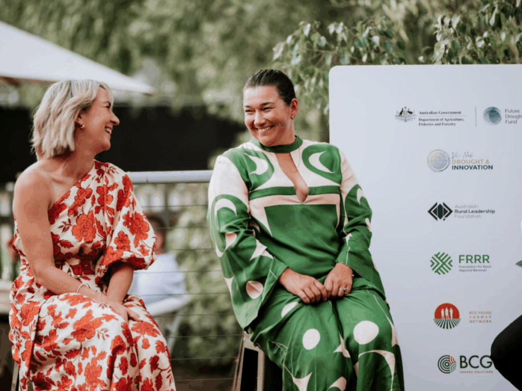 Two formally dressed women are seated on stools and the shot captures them smiling at each other. 