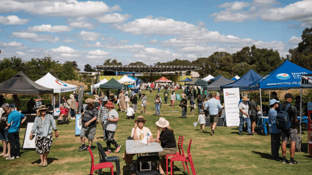 People participating in a field day in a way they might through a community benefit fund. There are marquees in the background and along both sides, with people walking around and some sitting at a table and chairs in the foreground.