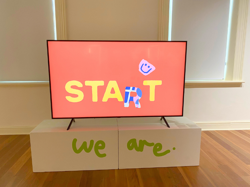 A TV sits on a white box with the writing 'We Are' on it, displaying a screen with a red background and the word start on it.