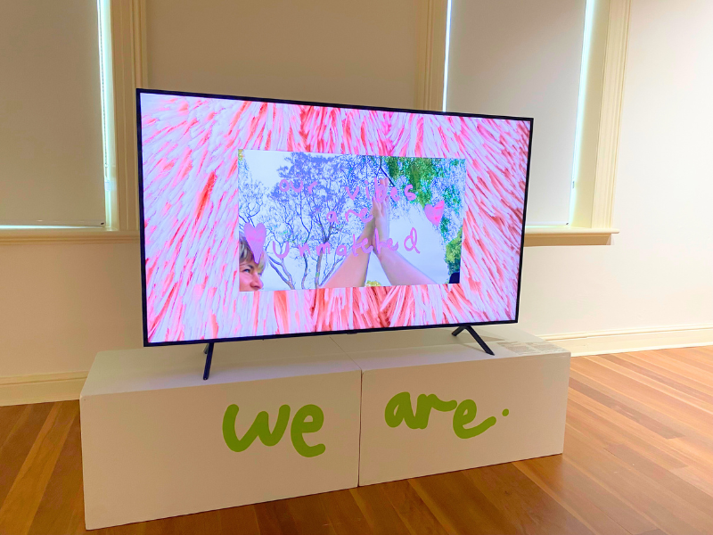 A TV sits on a white box with the writing 'We Are' on it, displaying pink imagery.