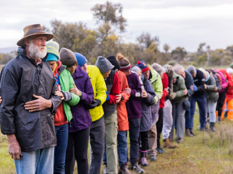 A group of people are in a line hugging each other like dominoes, wearing colourful clothing.