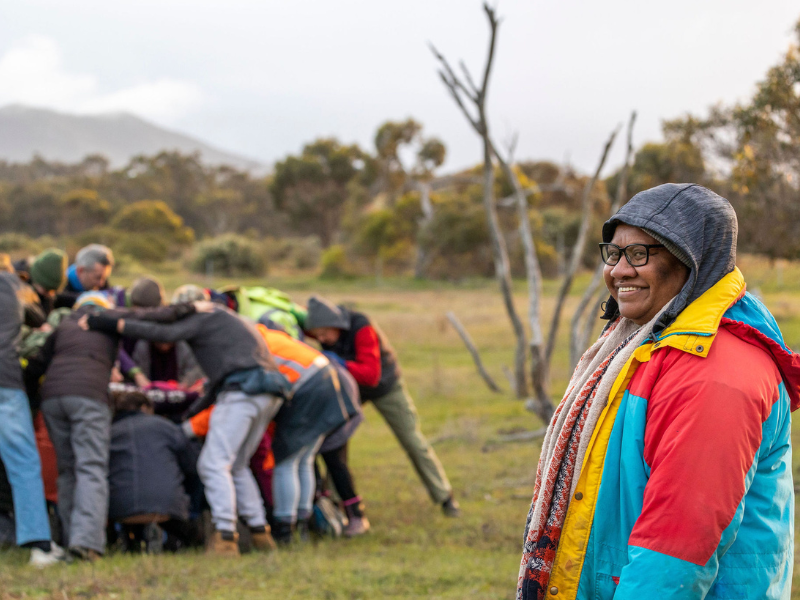 In the foreground a woman is looking beyond the camera and smiling, while a group of people are huddled in a scrum behind her.