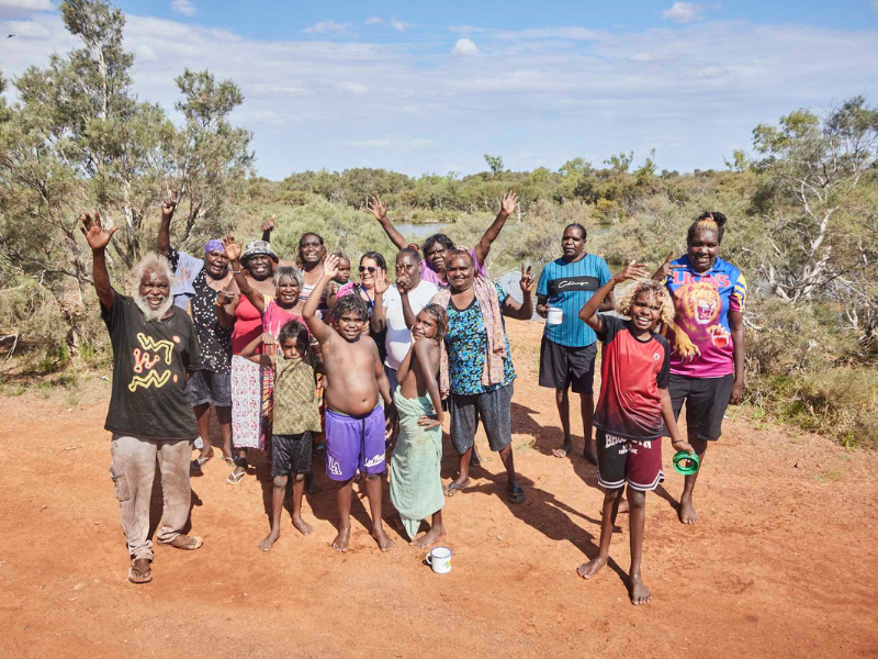 Group of young people are waving, standing together out in a remote setting with red dirt, greenery and a blue sky behind them.