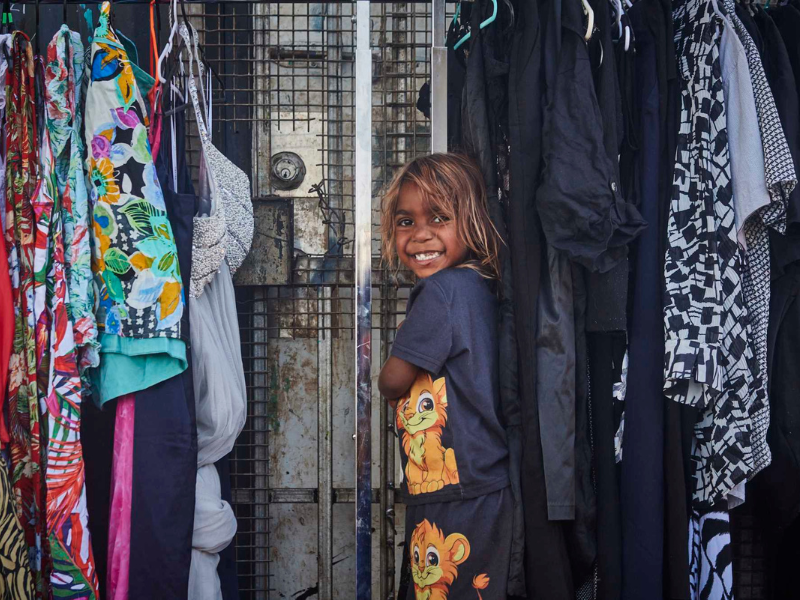 A young girl smiles at the camera in front of a collection of clothes handing. 