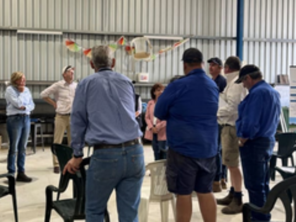 Farmers gathered together at a community event. Some men have their back to the camera, leaning on a chair with their arms.