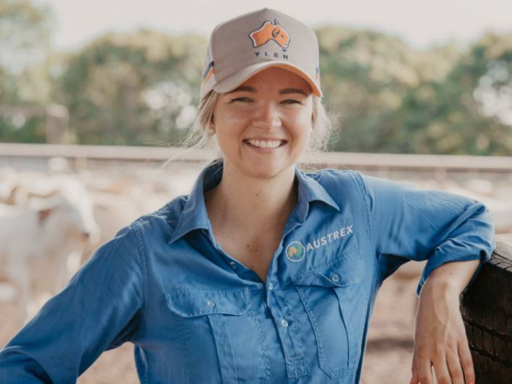 Young woman leaning on a fence in cattle yards. She's wearing a cap and there are cattle behind her.