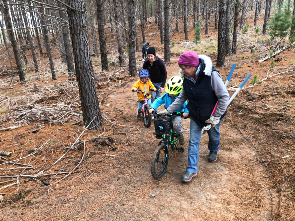 Two adult women assist two young children to ride bikes through a trail in the woods.