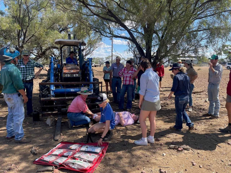 Group of people gathered in a mock first aid scenario outdoors near a tractor. 