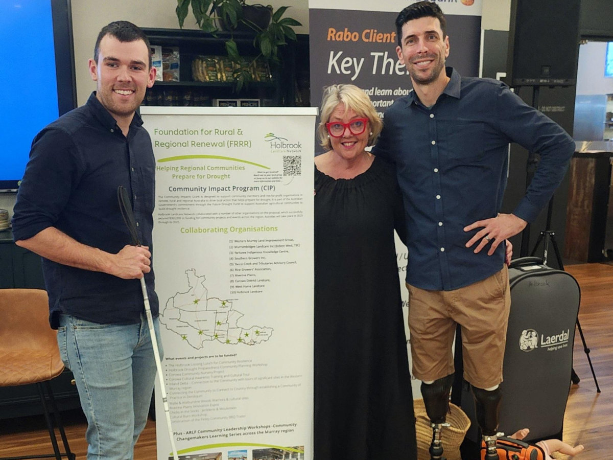 A woman stands between two taller men, one holding a mobility cane, the other with prosthetic legs. The three of them stand with a brand board. 