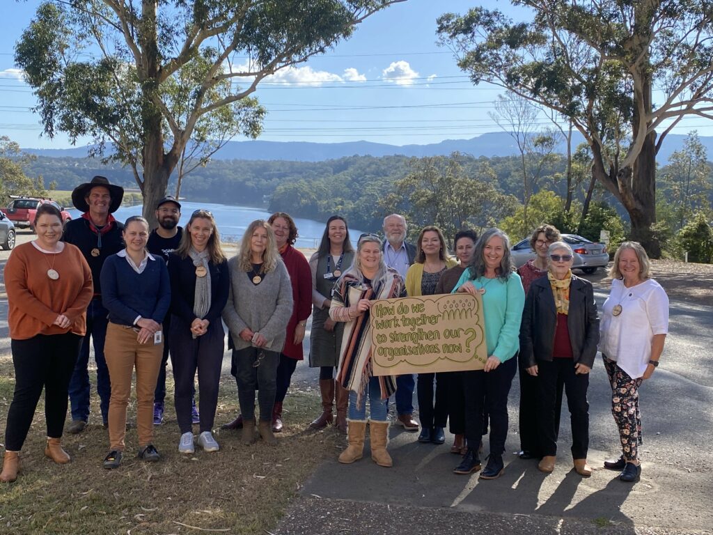 A group of people standing in front of a dam, holding a sign that says "How do we work together to strengthen our organisations now?"