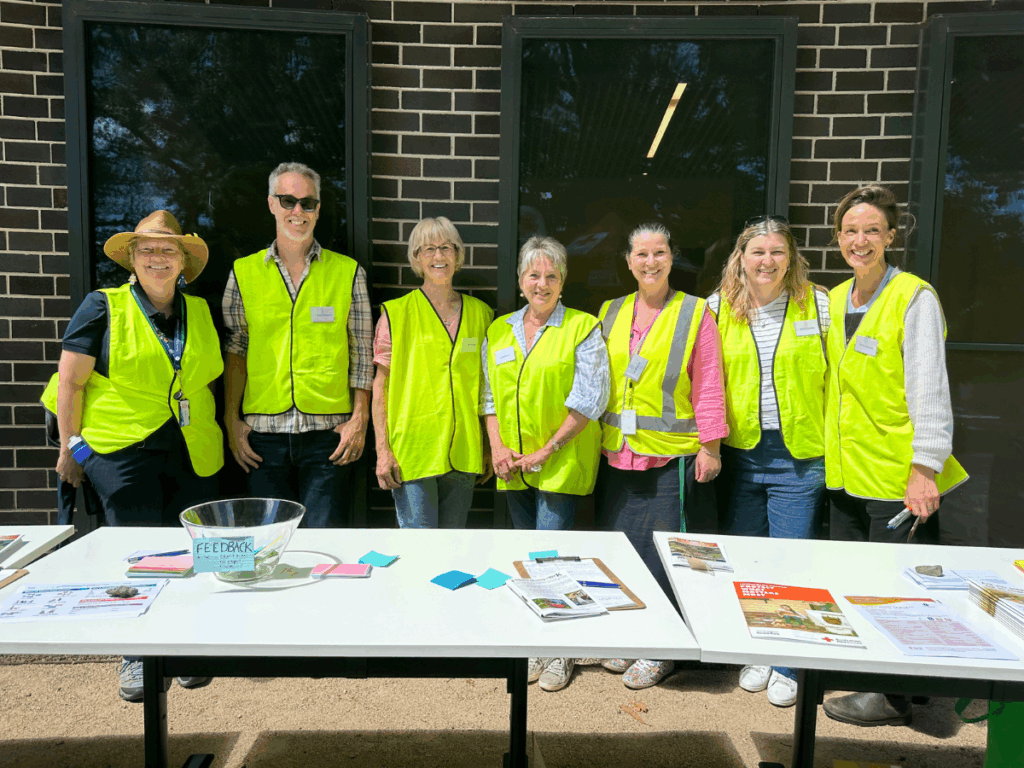 7 people are standing in a line wearing high-vis jackets in front of a pop-up table. 