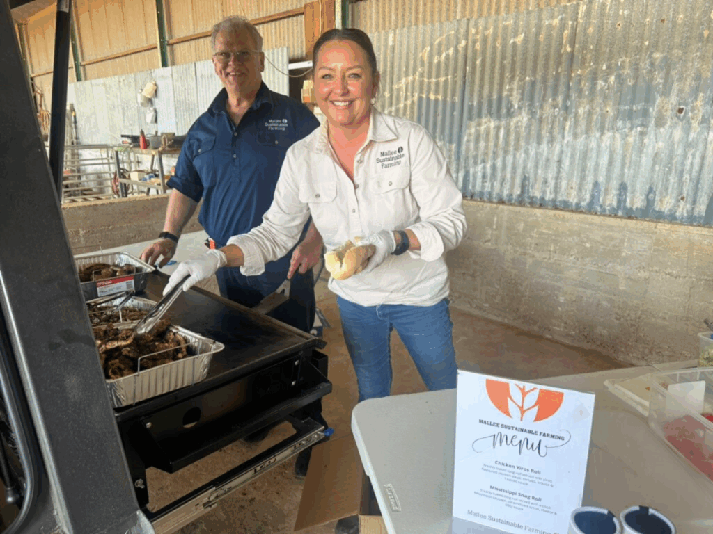 A brunette woman is cooking meat on a BBQ with a man standing behind her right shoulder. 