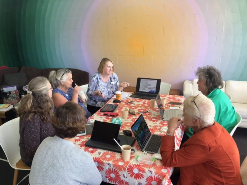 Group of women sit around a table with laptops. The wall in the background has a colorful light display of oranges, pinks and greens.