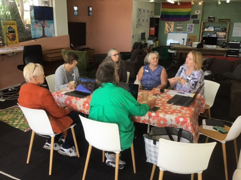 Older women gather at a table in a cafe setting talking to each other. 