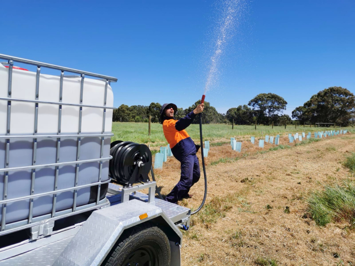 Male volunteer in an orange and navy jacket stands in a crop field with a water hose, aiming it at the sky. 
