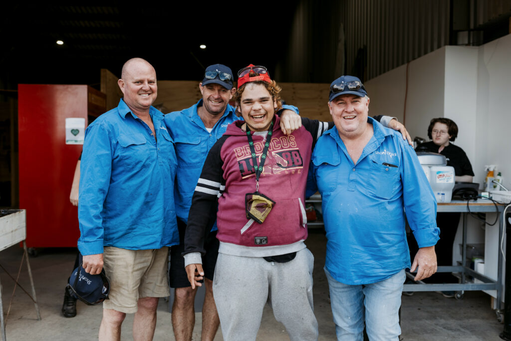 A group of four people standing with their arms around each other. There is a person sitting at a desk behind them.