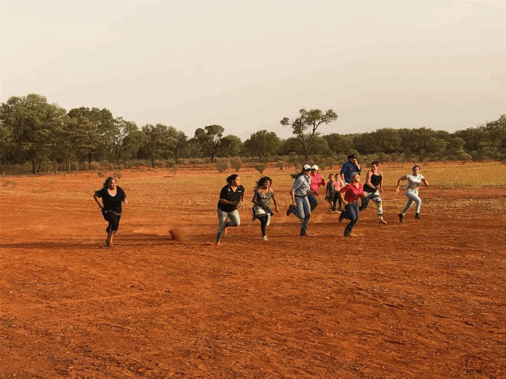 Group of kids are running in an open field made up of red dirt. 