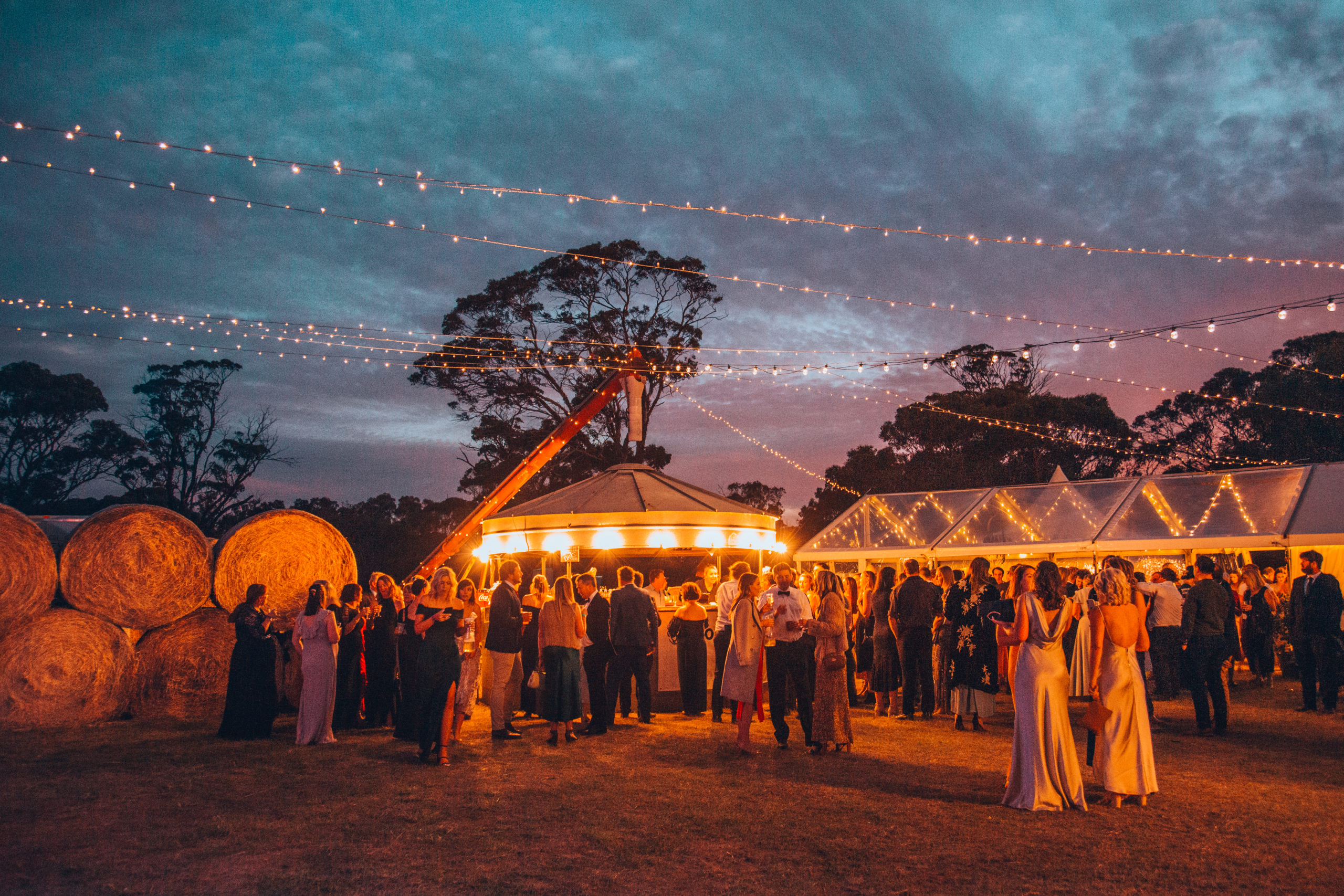 A crowd in evening wear under fairylights, surrounded by gum trees and hay bales.