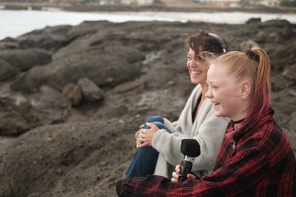 Two young women sitting on rocks by the water, one of them holds a microphone, both are smiling.