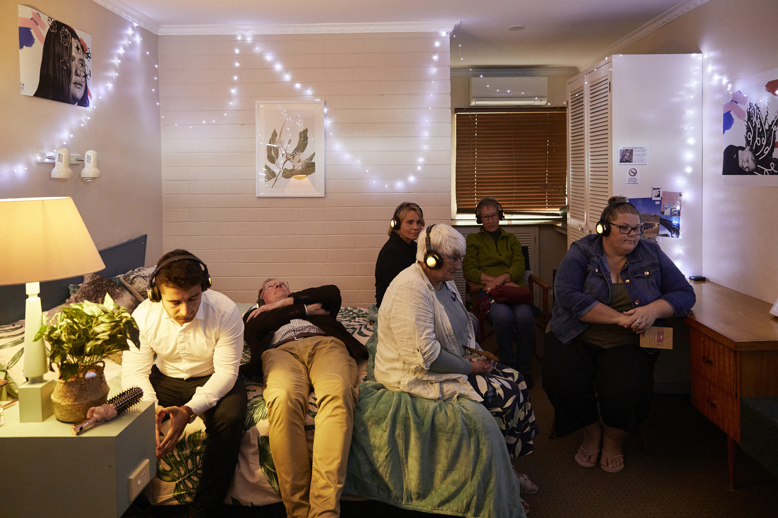 People sit on a bed and furniture in a bedroom with headphones on, listening to recordings.
