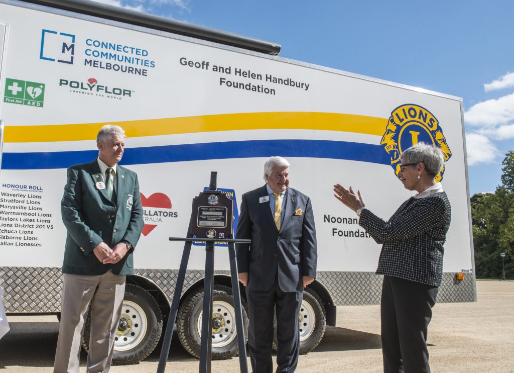 A woman claps and two men stand next to a plaque at an official event for the Lions Club mobile cancer screening unit