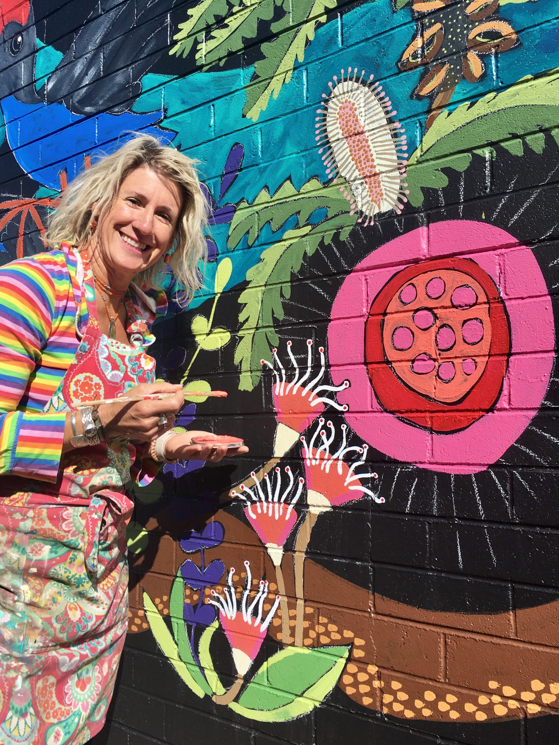 A woman in a rainbow striped shirt in the midst of painting a colourful mural of flora and fauna.