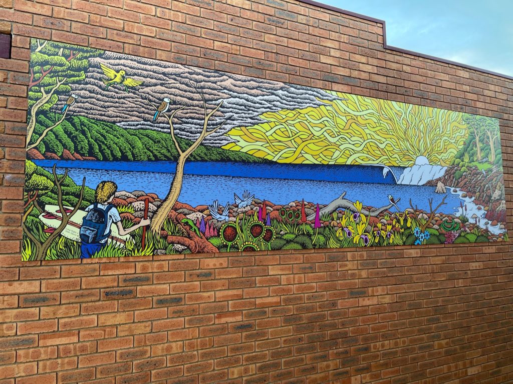A mural of a stylised coastal scene with big skies, colourful flora and birdlife and a boy walking holding a surfboard.