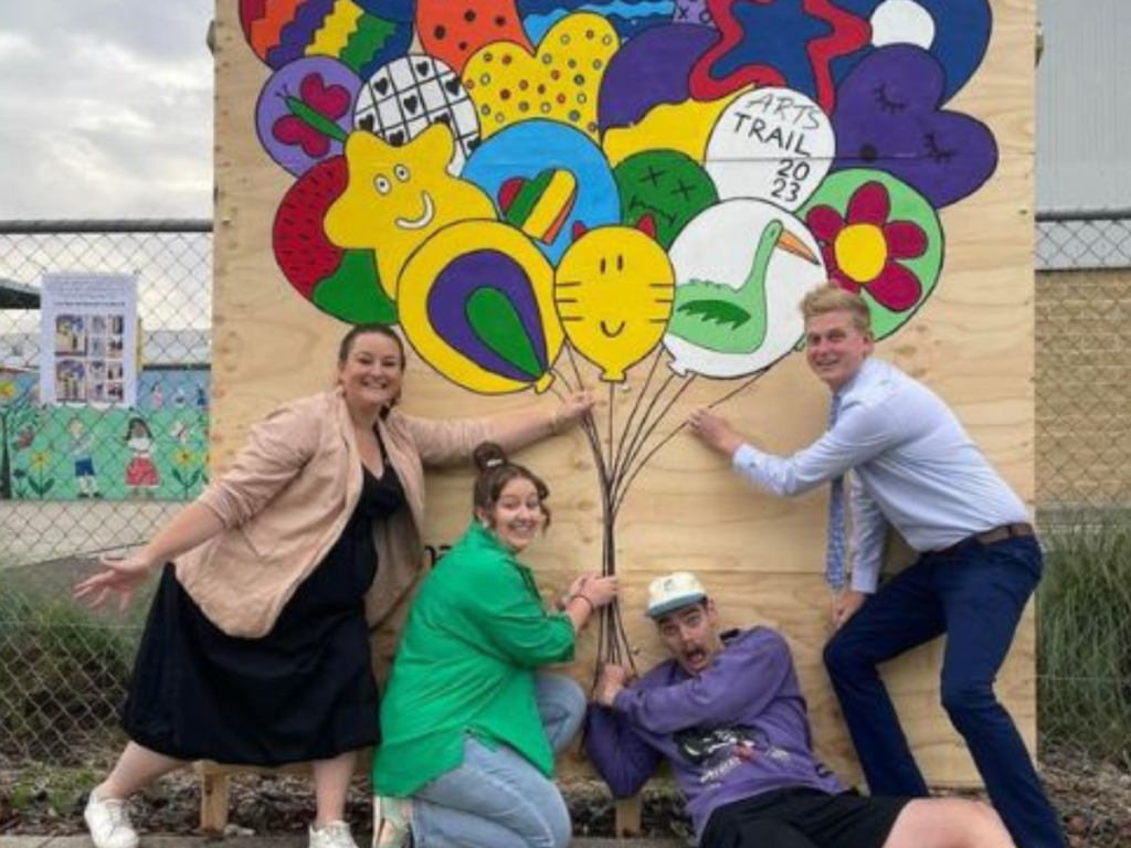 Four young adults in front of wall with a mural of a bunch of balloons painted on it.