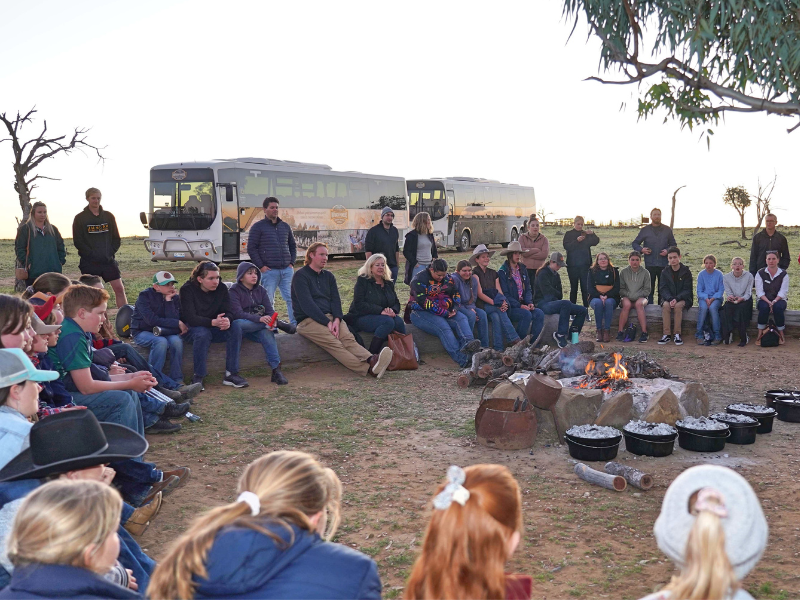Lots of people sitting in a circle around a campfire, with two buses and a sunset in the background.