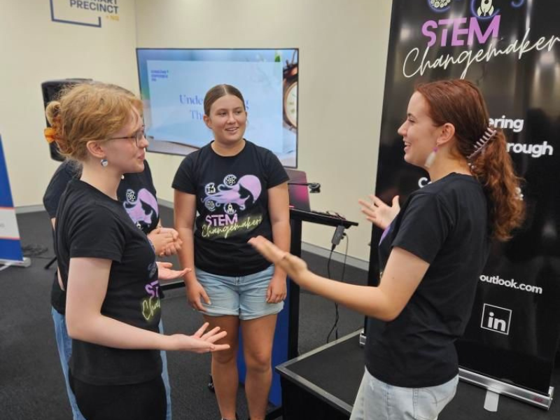 Three girls stand looking at each other smiling and gesturing with hands. 