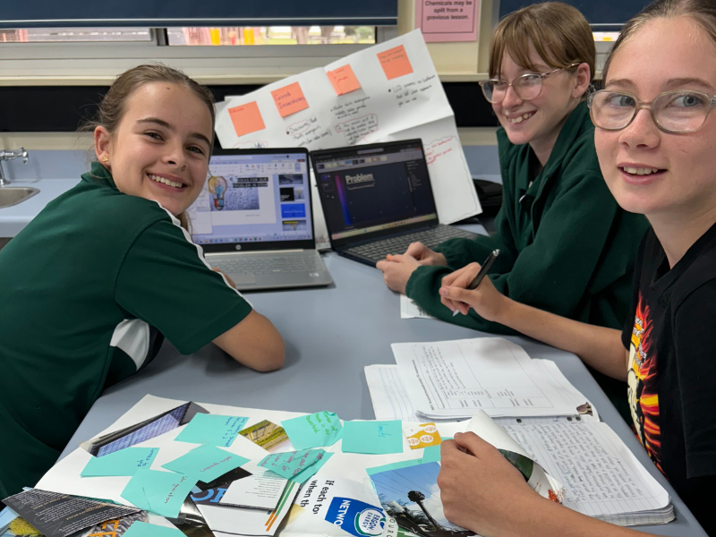 Three girls smile at the camera whilst working at a table with papers and a laptop. 
