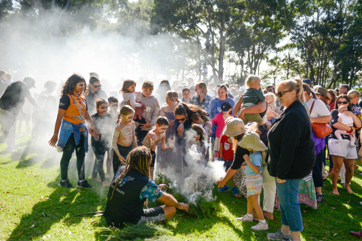 Children and adults gathered around a person crouched down who is performing a traditional Aboriginal smoking ceremony.