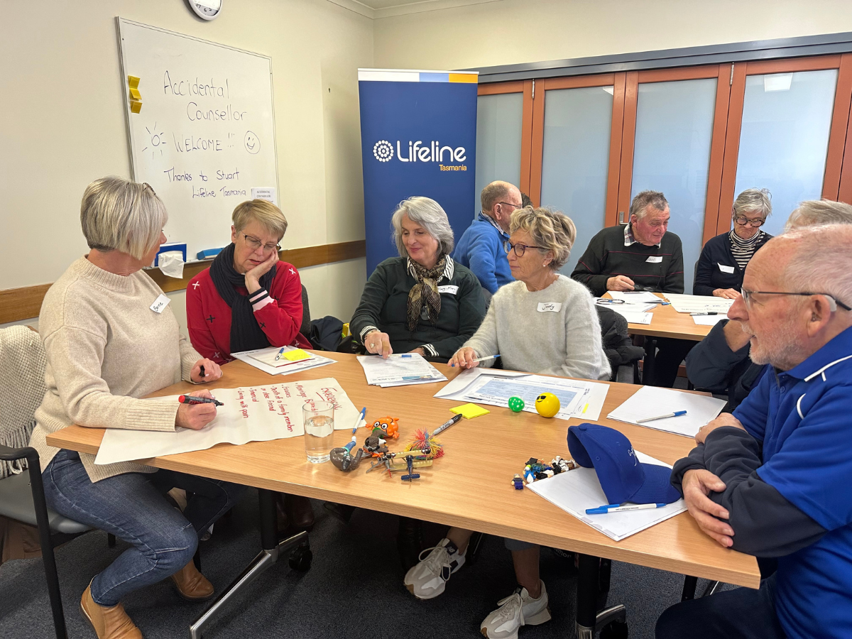 Group of older people are seated at a table writing and drawing on various pieces of paper. Another group sits beside them.