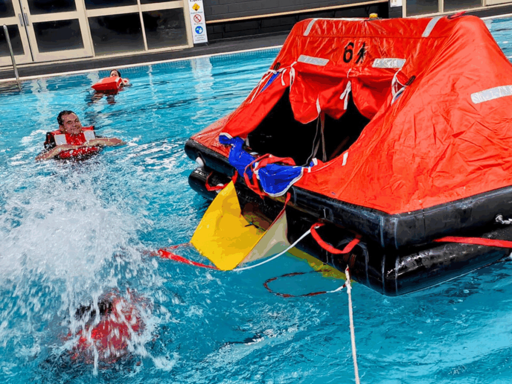 Two people in red life jackets are in a pool with a red and black inflatable, completing training.