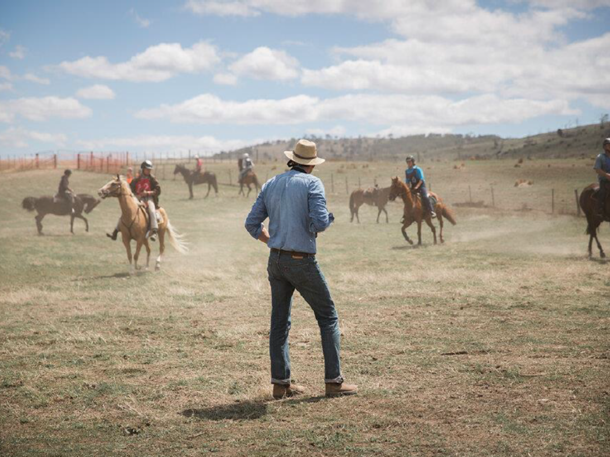 Man in cowboy hat stands in a dry field watching people riding horses. 