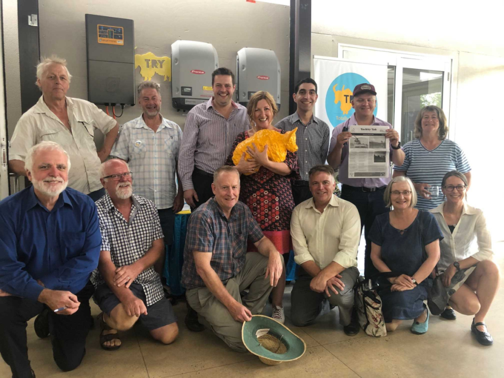 Group of people in two rows, with the ones in front crouched down. Behind them is a row of solar batteries.