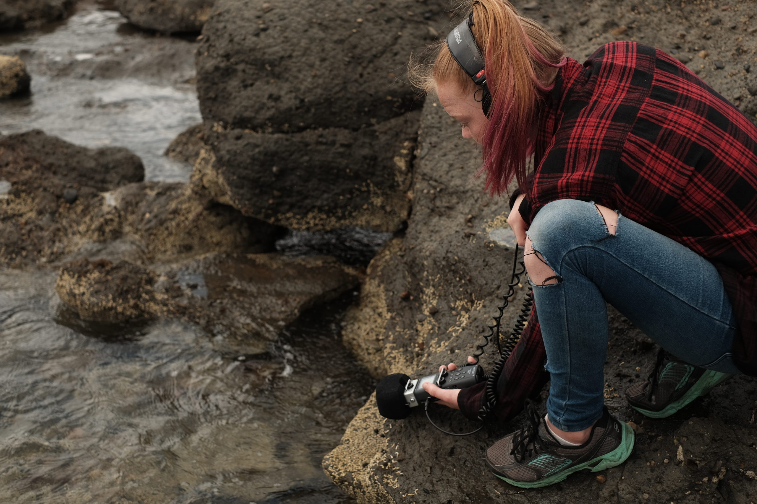 A young woman sitting on a rock by the water, recording sounds for a podcast