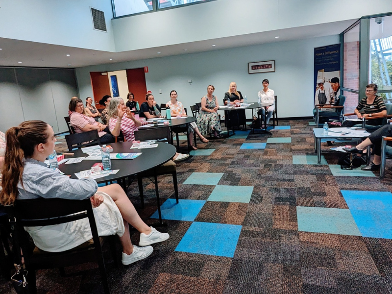 Classroom setting with a group of women sitting down at tables with light blue walls and brown and blue checkered carpert.  