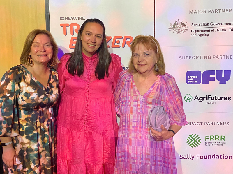 Three women standing together in front of a media board.