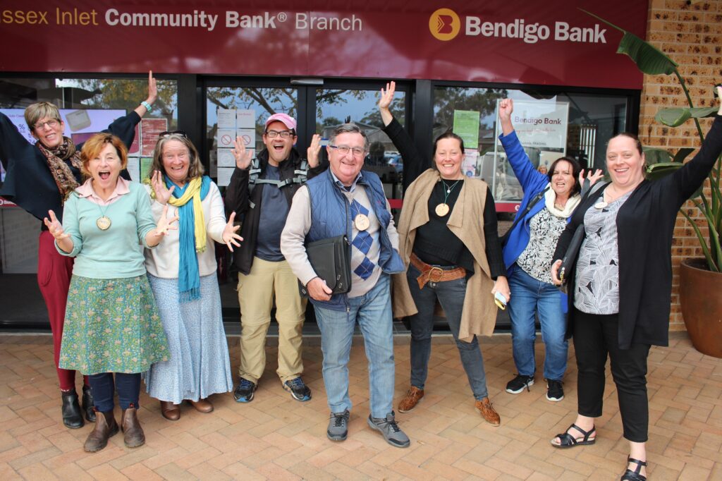 A group of people standing together and cheering in front of the Sussex Inlet branch of the Bendigo Bank.