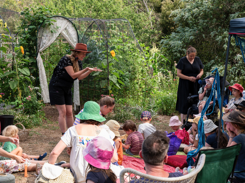 Group of people with lots of kids sitting on the  ground and a garden in the background of the photo.
