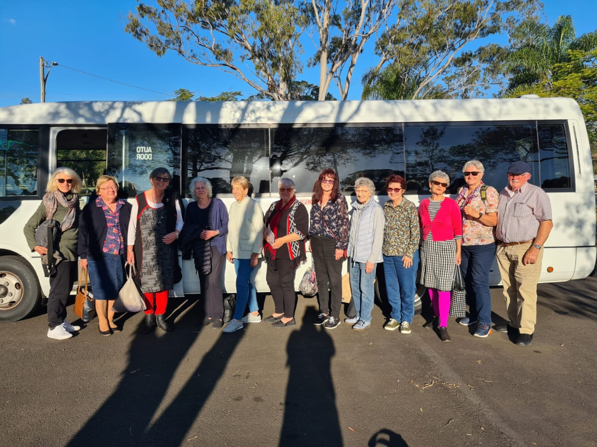 Group of people stand in front of a bus on tarmac for photo. Blue sky and colorful clothing. 